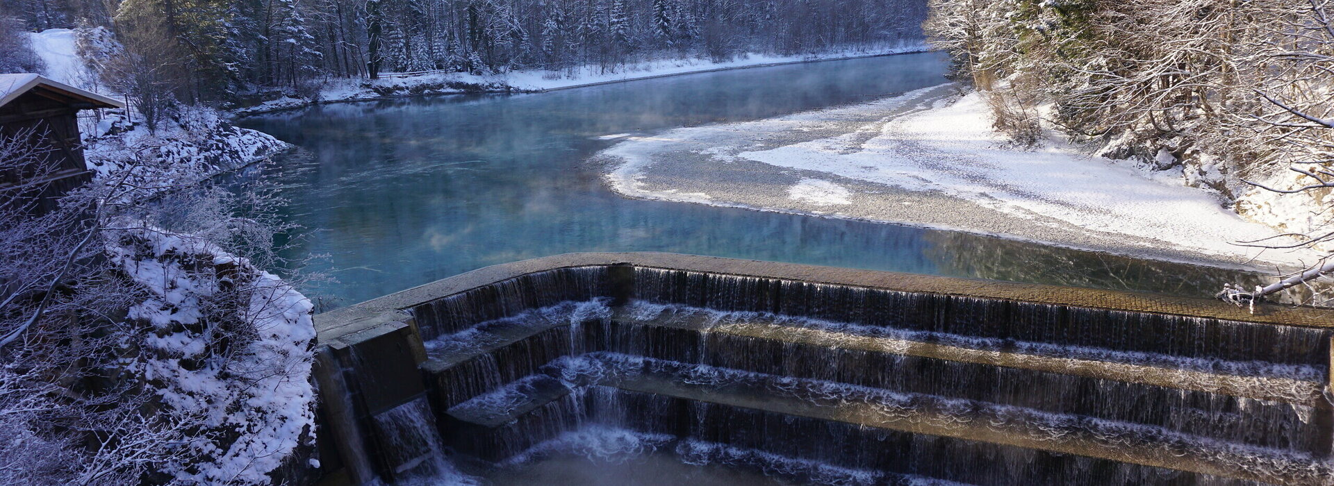 Das Bild zeigt eine friedliche Winterlandschaft. Ein dunkelblauer Fluss schlängelt sich durch die Landschaft. Der Fluss wird von einem schneebedeckten Wald flankiert, dessen Bäume sich gegen den Himmel abheben. Der Fluss fließt über gemauerte Steinstufen hinab. Der Himmel ist klar und blau, was einen wunderschönen Kontrast zum verschneiten Panorama bildet. Das Bild wird von einem hohen Standpunkt aus aufgenommen, der einen Panoramablick auf den Fluss und seine Umgebung bietet. 