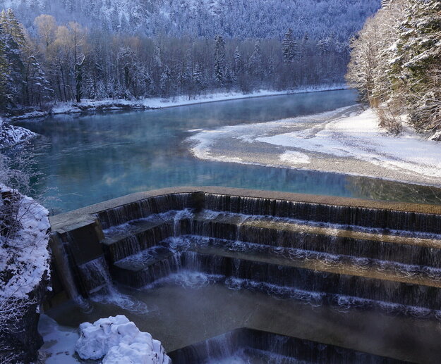 Das Bild zeigt eine friedliche Winterlandschaft. Ein dunkelblauer Fluss schlängelt sich durch die Landschaft. Der Fluss wird von einem schneebedeckten Wald flankiert, dessen Bäume sich gegen den Himmel abheben. Der Fluss fließt über gemauerte Steinstufen hinab. Der Himmel ist klar und blau, was einen wunderschönen Kontrast zum verschneiten Panorama bildet. Das Bild wird von einem hohen Standpunkt aus aufgenommen, der einen Panoramablick auf den Fluss und seine Umgebung bietet. 