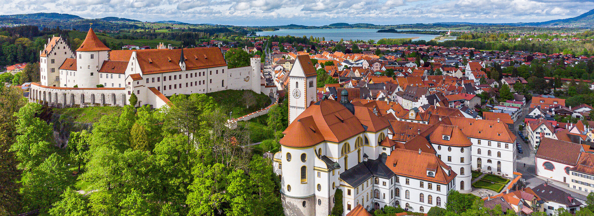Die Abbildung zeigt eine malerische Stadt mit einer Burgenstruktur, dem Hohen Schloss und auf das alte Kloster St. Mang, die von reichlich grünen Bäumen umgeben ist. Die Stadt ist in einem Tal gelegen, mit einem Fluss, der durch sie hindurchfließt. Im Hintergrund sieht man den Forggensee.