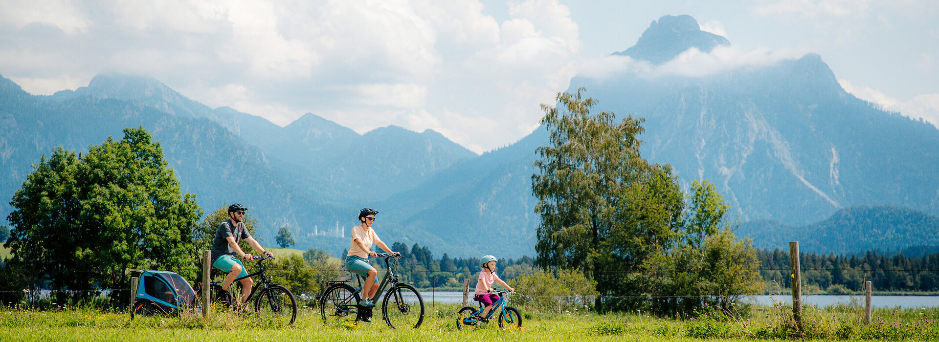 Das Bild fängt eine ruhige Szene auf einer grünen Wiese ein, wo drei Personen eine Fahrradtour genießen. Die Wiese ist üppig und grün, sie bietet ein lebhaftes Hintergrundbild zur Szene. Der Himmel darüber ist ein klarer Blau, der mit flauschigen weißen Wolken übersät ist. In der Ferne heben sich majestätische Berge gegen den Himmel ab und verleihen der Landschaft ein Gefühl von Größe. Die Personen in der Szene tragen legere Kleidung, was auf einen entspannten Tag hindeutet. Die Fahrräder, die sie fahren, sind deutlich zu erkennen, wobei eine Person ein Fahrrad fährt und die anderen zwei Fußgänger sind. Die Gesamtatmosphäre des Bildes ist eine von Ruhe und Naturgenuss.