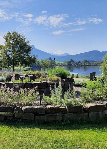 Das Bild zeigt eine große Kräuterspirale, auf der viele unterschiedliche Kräuter wachsen, die teilweise blühen. Im Hintergrund ist ein See zu sehen. Dahinter erheben sich Berge und bewaldete Hügel. Der Himmel ist blau mit weißen Wolken. Die Sonne scheint.