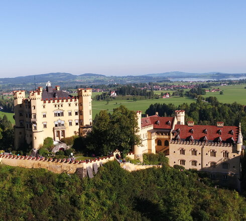 Das Bild zeigt das gelbe Schloss Hohenschwangau, dass auf einer Anhöhe thront und von Bäumen umgeben ist. Das Schloss besteht aus zwei Gebäudeteilen. Der quadratische, vierstöckige Hauptbau ist an jeder Ecke mit einem Turm versehen, der bis zum Dach reicht. Das längliche Nebengebäude steht mit ein bisschen Abstand zum Hauptgebäude.  Das Hauptgebäude ist von einer Mauer umgeben. An den Fenstern des Hauptgebäudes sind weiß, blaue Jalousien angebracht. Auf dem Dach des Hauptgebäudes steht eins überlebensgroße, weiße Schwanenfigur. In der Ferne sieht man eine Ortschaft und einen großen, blauen See. Der Himmel ist blau. Die Sonne scheint.