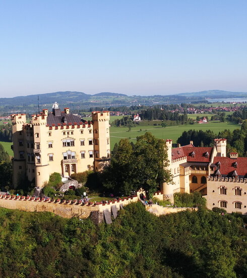 Das Bild zeigt das gelbe Schloss Hohenschwangau, dass auf einer Anhöhe thront und von Bäumen umgeben ist. Das Schloss besteht aus zwei Gebäudeteilen. Der quadratische, vierstöckige Hauptbau ist an jeder Ecke mit einem Turm versehen, der bis zum Dach reicht. Das längliche Nebengebäude steht mit ein bisschen Abstand zum Hauptgebäude.  Das Hauptgebäude ist von einer Mauer umgeben. An den Fenstern des Hauptgebäudes sind weiß, blaue Jalousien angebracht. Auf dem Dach des Hauptgebäudes steht eins überlebensgroße, weiße Schwanenfigur. In der Ferne sieht man eine Ortschaft und einen großen, blauen See. Der Himmel ist blau. Die Sonne scheint.