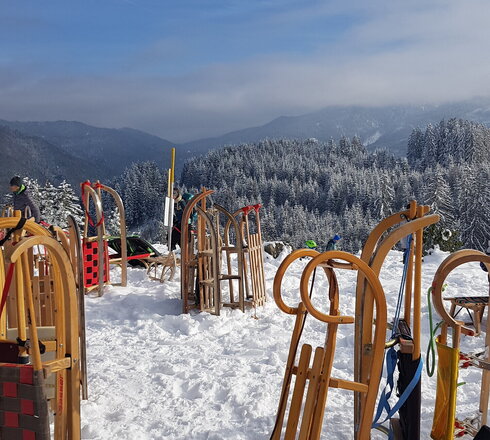 Das Bild fängt eine friedliche Winterlandschaft ein. Der Boden ist von einer dicken Schneeschicht bedeckt, die den klaren blauen Himmel darüber widerspiegelt. Verstreut in der verschneiten Landschaft stehen hölzerne Strukturen, deren natürliches Braun sich vom weißen Schnee abhebt. Diese Strukturen scheinen Spielgeräte zu sein, einschließlich Schaukeln und Rutschen, die auf einen Ort hinweisen, an dem Kinder die Winterlandschaft erkunden und genießen können. Die Geräte werden gerade nicht genutzt, was durch die Abwesenheit von Menschen auf dem Bild bestätigt wird. Die Gesamtstimmung vermittelt ein Gefühl von Ruhe und den einfachen Freuden des Winterspiels.