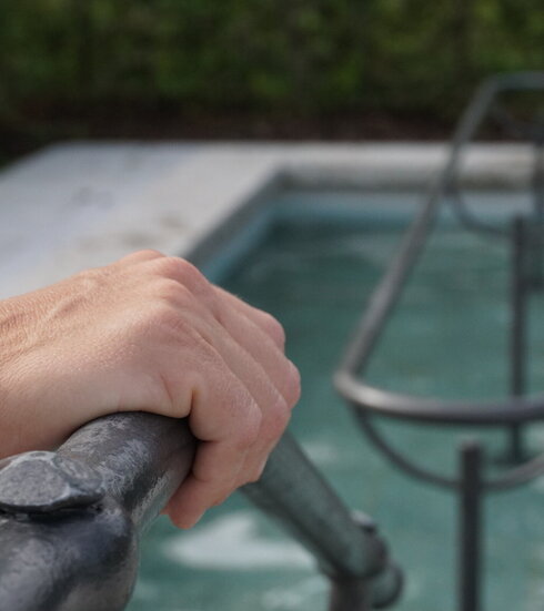 Das Bild zeigt eine Hand, die sich auf einem Metallgeländer abstützt, das in eine Kneipp-Tretbecken führt. Innerhalb des Tretbeckens verläuft ein Geländer. Das Tretbecken ist mit Wasser gefüllt.       