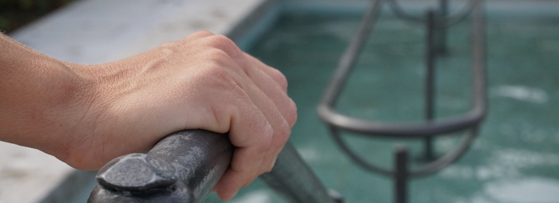 Das Bild zeigt eine Hand, die sich auf einem Metallgeländer abstützt, das in eine Kneipp-Tretbecken führt. Innerhalb des Tretbeckens verläuft ein Geländer. Das Tretbecken ist mit Wasser gefüllt.       