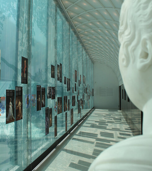 Man blickt einen langen Gang entlang. Die Decke besteht aus rautenförmigen Kassetten. Auf der linken Seite des Bildes befindet sich eine hohe, fast durchsichtige Wand, die mit Blattwerk und Ästen durchzogen ist.  Viele Bildnisse historischer Persönlichkeiten sind zu sehen. Im Vordergrund ist das Profil einer weiblichen Marmorbüste zu sehen. Die ebenfalls scheinbar den Gang entlang blickt. Man schaut ihr über die Schulter. 