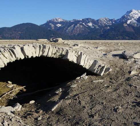 Das Bild zeigt ein steinernes Gewölbe, das in eine bergige Landschaft eingebettet ist. Das Gewölbe ist der Mittelpunkt des Bildes und erinnert an den gemauerten Unterbau einer alten Brücke. Das Gewölbe ist von einem steinigen, tristen und kargen Terrain umgeben, was der Szene einen rauen Charme verleiht. Die Berge im Hintergrund sind teilweise mit Schnee bedeckt. Der Himmel darüber ist klar und blau. Es handelt sich um den Seegrund des leeren Stausees Forggensee, der die Mauern der Dörfer freigibt, die sich hier vor dem See befunden haben..