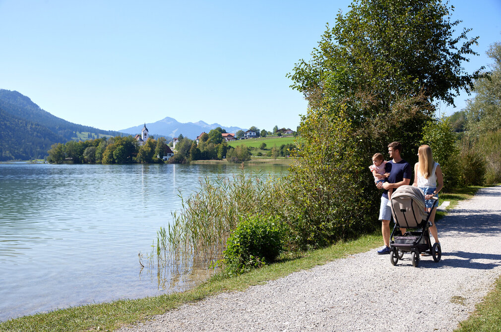Das Bild zeigt eine friedliche Szene einer Familie von vier Personen, die entlang eines Schotterwegs an einem See spazieren. Der Vater, der einen weißen Hemd trägt, schiebt einen Kinderwagen, während die Mutter, die ein blaues Hemd trägt, neben ihm geht. Ihre beiden Kinder, ein Junge und ein Mädchen, sind ebenfalls Teil der Familienausflug. Der See, der ruhig und einladend ist, liegt eingebettet in üppig grünen Bäumen und Bergen in der Ferne. Die Familie scheint einen gemütlichen Spaziergang zu genießen, während sie die natürliche Schönheit um sie herum aufnehmen.