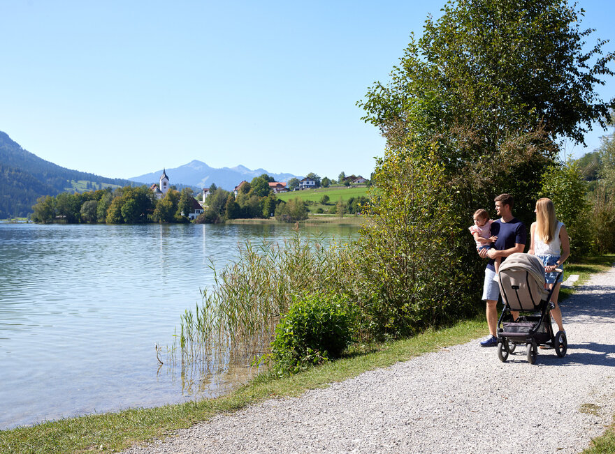 Das Bild zeigt eine friedliche Szene einer Familie von vier Personen, die entlang eines Schotterwegs an einem See spazieren. Der Vater, der einen weißen Hemd trägt, schiebt einen Kinderwagen, während die Mutter, die ein blaues Hemd trägt, neben ihm geht. Ihre beiden Kinder, ein Junge und ein Mädchen, sind ebenfalls Teil der Familienausflug. Der See, der ruhig und einladend ist, liegt eingebettet in üppig grünen Bäumen und Bergen in der Ferne. Die Familie scheint einen gemütlichen Spaziergang zu genießen, während sie die natürliche Schönheit um sie herum aufnehmen.