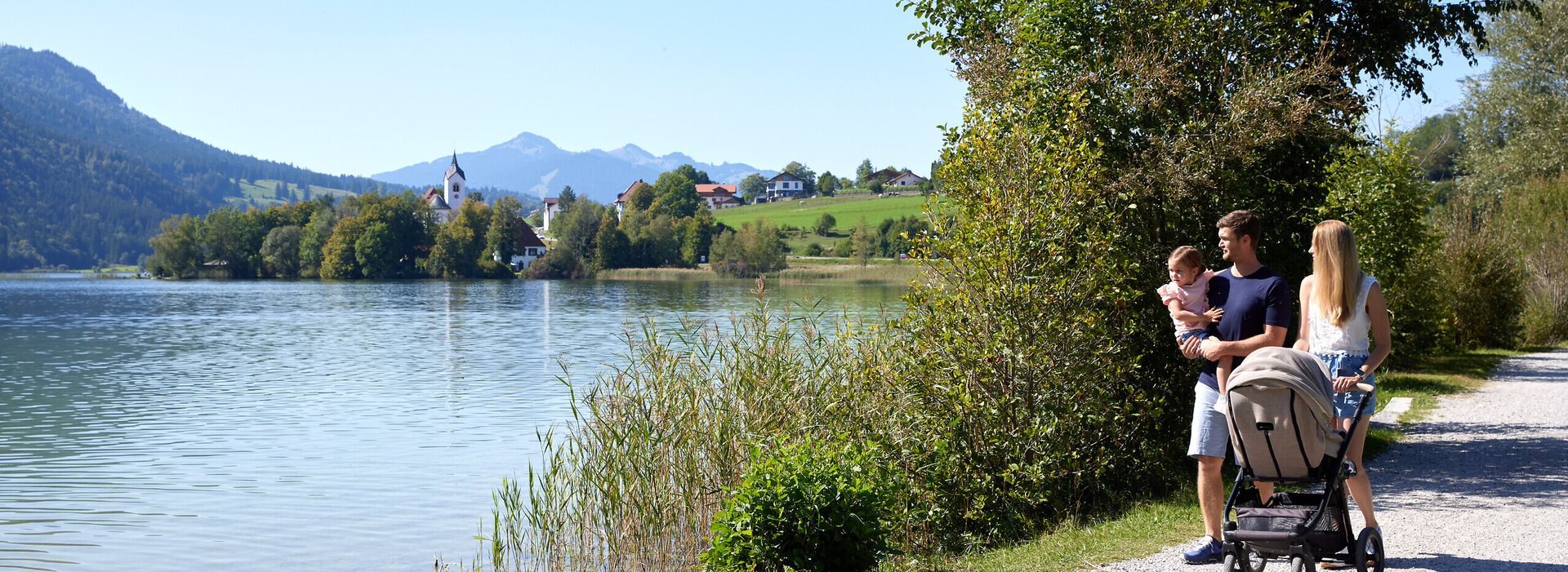 Das Bild zeigt eine friedliche Szene einer Familie von vier Personen, die entlang eines Schotterwegs an einem See spazieren. Der Vater, der einen weißen Hemd trägt, schiebt einen Kinderwagen, während die Mutter, die ein blaues Hemd trägt, neben ihm geht. Ihre beiden Kinder, ein Junge und ein Mädchen, sind ebenfalls Teil der Familienausflug. Der See, der ruhig und einladend ist, liegt eingebettet in üppig grünen Bäumen und Bergen in der Ferne. Die Familie scheint einen gemütlichen Spaziergang zu genießen, während sie die natürliche Schönheit um sie herum aufnehmen.