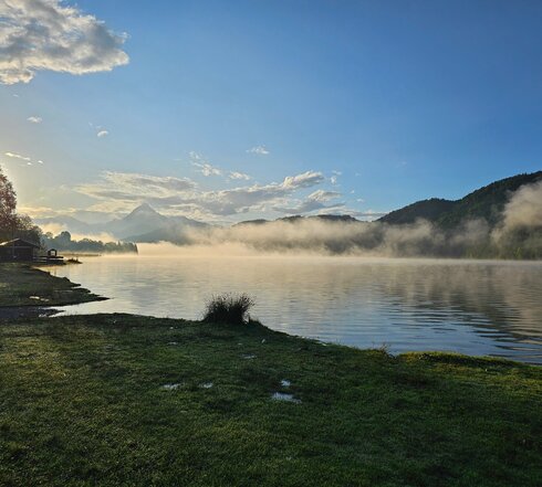 Das Bild zeigt einen See im Morgenlicht. Über dem See liegen dichte Nebelschwaden, durch die sich die Sonne ihren Weg bahnt. Der Himmel ist blau. Das Ufer im Vordergrund ist mit Gras bewachsen. Im Hintergrund erheben sich grüne Hügel. 