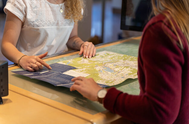 Das Bild zeigt eine Karte, die auf einem Tresen liegt. Auf beiden Seiten des Tresens sind Personen zu sehen, die die Karte betrachten. Man sieht nur Teile der Arme und Oberkörper der Frauen. Eine Person zeigt etwas auf der Karte. 