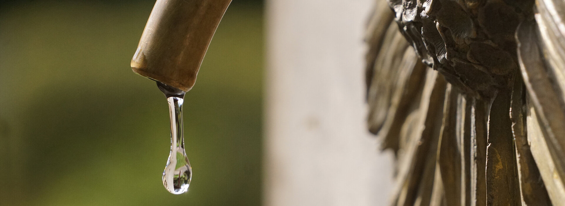 Zu sehen ist die Nahaufnahme eines messingfarbenen Wasserhahns, aus dem ein einzelner Wassertropfen herab perlt. Der Wasserhahn kommt aus einer messingfarbenen Arnikablüte, die an einem aufrecht stehenden Stein angebracht ist.