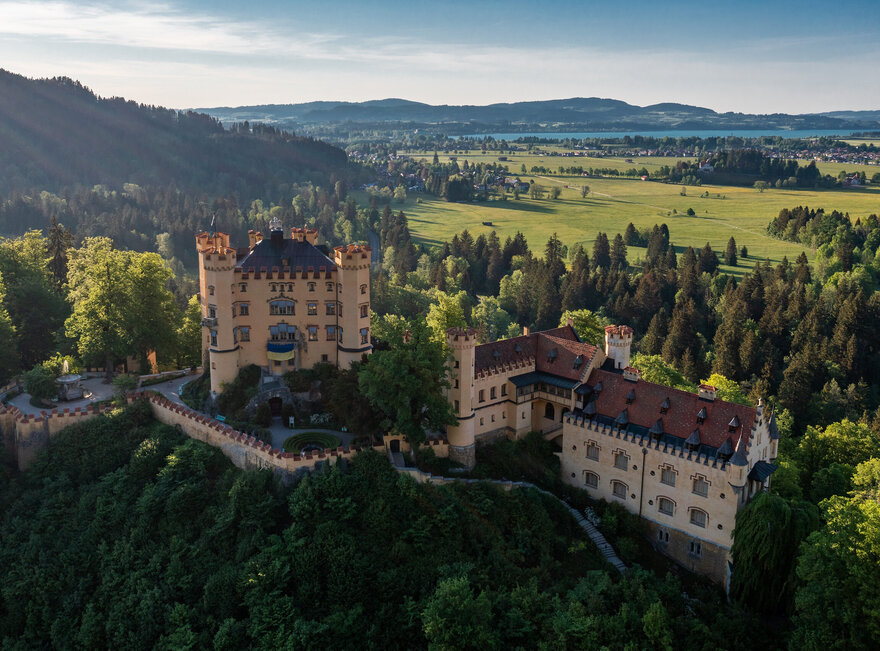 Das Bild zeigt das Schloss Hohenschwangau, das auf einem Hügel thront und von üppigem grünen Wald umgeben ist. Die Burg, aus Stein erbaut, verfügt über mehrere Türme und Erker, welche zu ihrer Größe beitragen. Die Architektur der Burg erinnert an eine mittelalterliche Festung, ausgestattet mit Wehrgängen und einem Graben. Die umliegende Landschaft ist gesprenkelt mit grünen Bäumen und sanften Hügeln, was eine malerische Kulisse schafft.