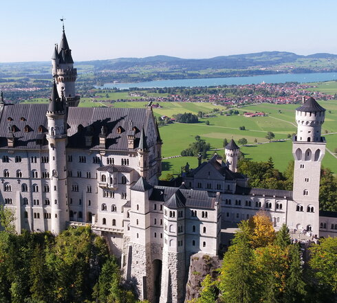 Das Bild zeigt das weiße Schloss Neuschwanstein, dass auf einer bewaldeten Anhöhe vor einer weiten Ebene liegt. Auf der Ebene sind Seen, die Gemeinde Schwangau und die Stadt Füssen zu sehen, die umgeben von Wäldern und grünen Wiesen sind. Das Schloss Neuschwanstein steht im Fokus des Fotos. Es besteht aus mehreren unterschiedlichen Gebäudeteilen, wie Türmen, Erkern und einem Torbau, die miteinander verbunden sind. Das Hauptgebäude des Schlosses ist 5-stöckig. Es hat ein graues Dach. Auf dem Hauptgebäude stehen eine überlebensgroße Statue sowie eine Löwenfigur. Der Himmel ist blau. Die Sonne scheint.