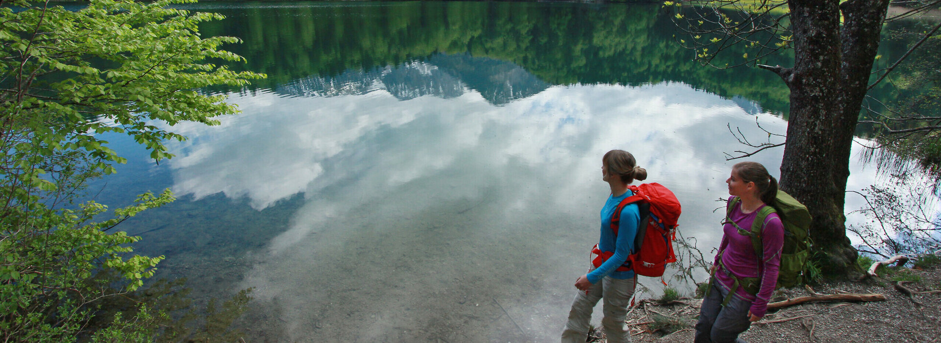 Auf dem Bild sieht man zwie Frauen mit Wanderkleidung und Wanderrucksäcken die an einem Seeufer laufen. Am Seeufer sind Bäumde und im Hintergrund in der Ferne schneebedkeckte Berge.