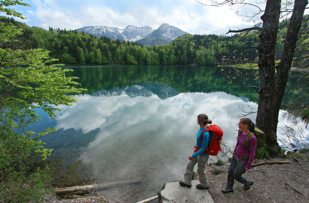 Auf dem Bild sieht man zwie Frauen mit Wanderkleidung und Wanderrucksäcken die an einem Seeufer laufen. Am Seeufer sind Bäumde und im Hintergrund in der Ferne schneebedkeckte Berge.