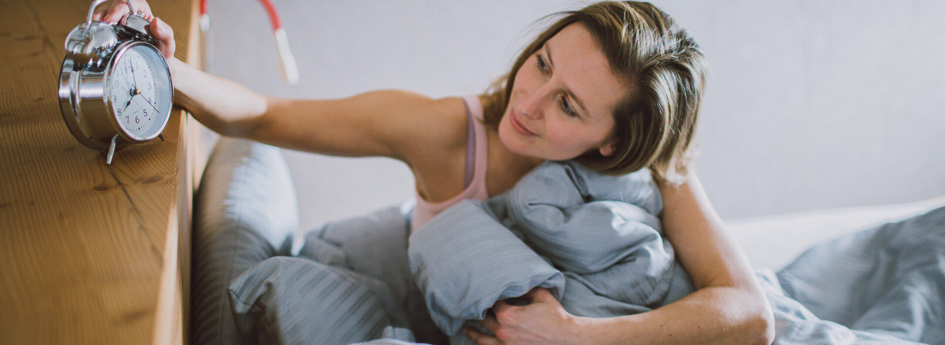 Das Bild zeigt eine Frau mit einem weißen kurzen Oberteil, die in einem hellgrau bezogenen Bett liegt. Sie hat ihren Oberkörper leicht angehoben und hält in ihre rechten Hand einen silbernen Wecker. Es sieht so aus als würde sie den Wecker ausschalten. 