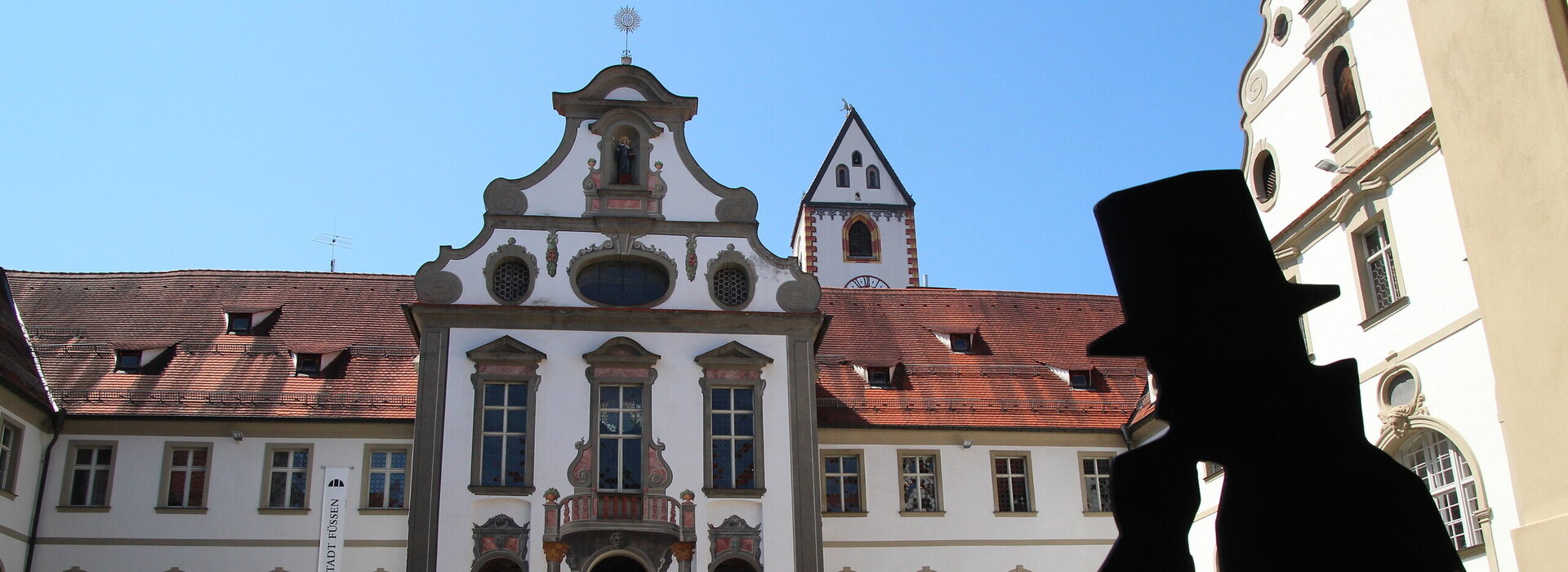 Das Foto zeigt den Klosterhof des ehemaligen Benediktinerklosters St. Mang. Der Blick fällt auf den Eingang des Museums der Stadt Füssen. Der Boden des Hofs ist mit Kopfsteinpflaster gepflastert. Die Fassade des Gebäudes ist mit Illusionsmalerei verziert. Vor dem Eingang des Museums liegt ein roter Teppich auf dem Boden. Der Himmel ist blau. Die Sonne scheint.