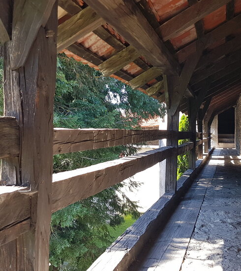 Das Bild zeigt einen überdachten Gang, der auf der einen Seite durch ein Holzgeländer begrenzt ist, auf der anderen Seite an eine historische Mauer grenzt. Die Sonne scheint.