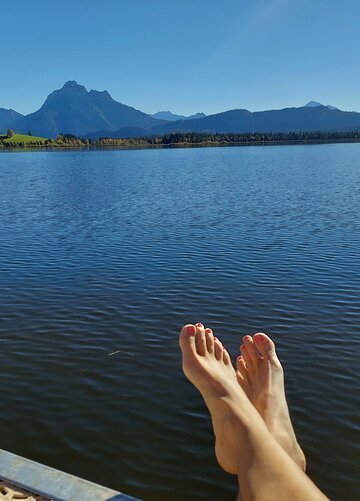 Das Bild zeigt einen See mit ruhiger Wasseroberfläche. Im Hintergrund erheben sich die Berge. Im Vordergrund sind zwei nackte Füße zu sehen, die wohl der Fotografin gehören. Die Fußnägel sind rot lackiert. Die Sonne scheint. Der Himmel ist blau. 