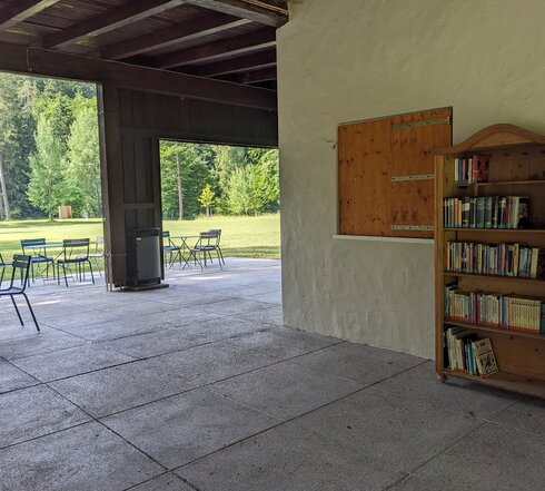 Das Bild ist unter einem großen Vordach aufgenommen. Der Boden ist mit Steinplatten ausgelegt, auf denen Tische und Stühle stehen. An der Hauswand lehnt ein geöffneter Bücherschrank, der mit Büchern gefüllt ist. Im Hintergrund beginnt ein Wald. Davor liegt eine grüne Wiese. Die Sonne scheint. 