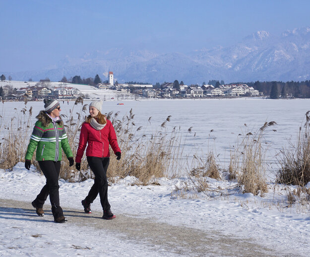 Das Bild fängt eine ruhige Winterlandschaft ein. Zwei Frauen sind auf einem schneebedeckten Uferweg zu sehen. Der Weg, auf dem sie gehen, ist von hohen Gräsern gesäumt. Der See ist zugefrohren. Im Hintergrund ist der Ort Hopfen zu erkennen. Jenseits der Ortschaft erheben sich majestätische Berge, deren Gipfel mit Schnee bedeckt sind. Der Himmel ist blau und sonnig. 