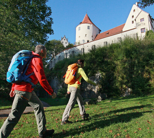 Das Bild zeigt einen Mann und eine Frau in Wanderkleidung und Rucksack, die eine Wiese entlang gehen. Über ihnen thront auf einem Felsen das Hohe Schloss. Im Hintergrund wachsen hohe grüne Bäume. Der Himmel ist blau. Die Sonne scheint.  