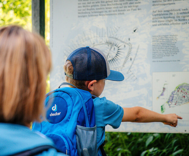  Das Bild zeigt einen Moment der Erkundung und des Lernens. Ein junger Junge, mit einem blauen Baseballcap und einer passenden blauen Schultertasche bekleidet, zeigt auf eine Karte auf einem Schild. Die Karte ist detailliert und zeigt verschiedene Orte und deren entsprechende Farben. Die Neugierde und das Interesse des Jungen an der Karte lassen darauf schließen, dass er sich mit den verschiedenen Orten und deren Bedeutung auseinandersetzt. Das Schild befindet sich in einem Park, was einen Kontext für die Erkundung des Jungen bietet. Die Parklandschaft in Verbindung mit dem Interesse des Jungen an der Karte zeichnet das Bild eines Tages im Freien, in dem er sich mit der Welt um ihn herum auseinandersetzt.