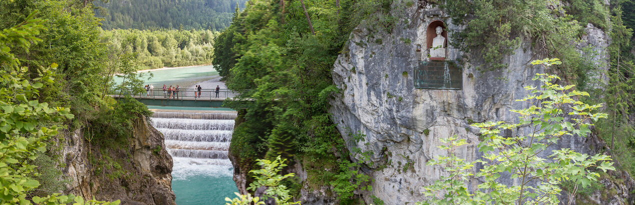 Das Bild fängt einen atemberaubenden Blick auf einen Wasserfall ein, der an einem felsigen Steilhang hinabstürzt. Der Wasserfall liegt im Fokus des Bildes. Das felsige Ufer ist von üppigem grünem Laub und Büschen geziert. Über die Schlucht, die der Wasserfall durchfließt, führt eine Brücke. In einen Felsabhang am Ufer ist eine Büste eingearbeitet. Sie zeigt König Max II. von Bayern. Die Perspektive des Bildes ist aus einer hohen Position, die auf den Wasserfall hinunterblickt, was die Szene zusätzlich in Größe erscheinen lässt.