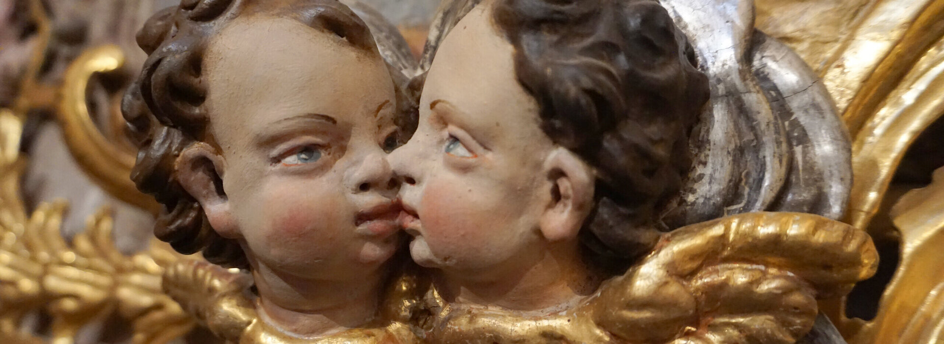 Das Foto zeigt die Köpfe zweier Putten, die sich an Mund und Nase berühren. Sich also scheinbar küssen. Die Puttenköpfe haben braune Haare und blaue Augen. An der Stelle der Kragen sind goldene Engelsflügel zu sehen. 