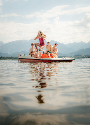 Das Bild zeigt eine Familie, einen Mann, eine Frau, ein Mädchen und einen Jungen. Sie sitzen auf einem Tretboot, dass inmitten eines Sees schwimmt. Alle tragen Badeklamotten. Das Mädchen hält sich die Nase zu und springt in den See. Im Hintergrund erheben sich die Berge. Der Himmel ist blau. Die Sonne scheint. 