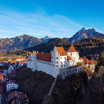 Man blickt von oben auf das Hohe Schloss, das auf einer Anhöhe über Füssen thront. Darunter sieht man die Häuser der Altstadt, die dicht aneinandergebaut wurden. Im Hintergrund erheben sich schneebedeckte Berge und bewaldete Hügel. Der Himmel ist blau. Die Sonne scheint. 
