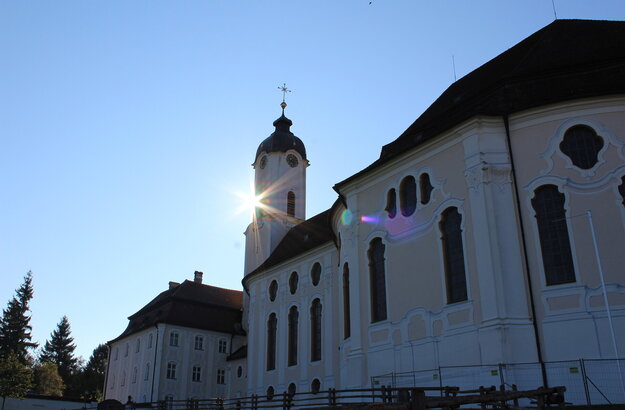  Das Bild zeigt die große weiße Wieskirche, mit einem hohen Kirchturm und einem Kreuz oben drauf. Die Sonne scheint hell auf die Kirche, sodass eine schöne und sommerliche Atmosphäre entsteht.