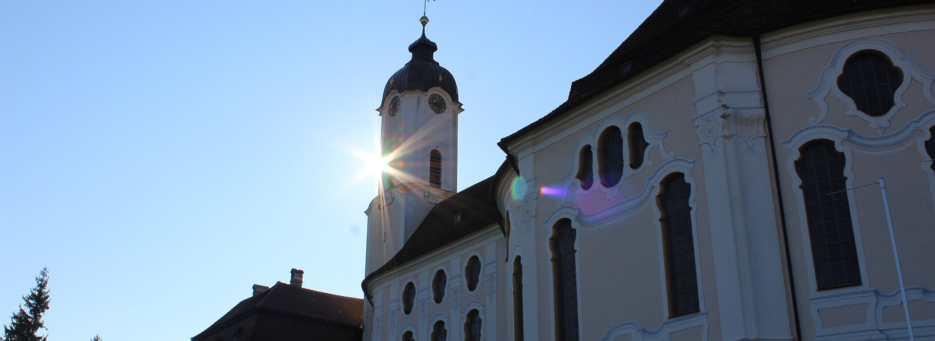  Das Bild zeigt die große weiße Wieskirche, mit einem hohen Kirchturm und einem Kreuz oben drauf. Die Sonne scheint hell auf die Kirche, sodass eine schöne und sommerliche Atmosphäre entsteht.