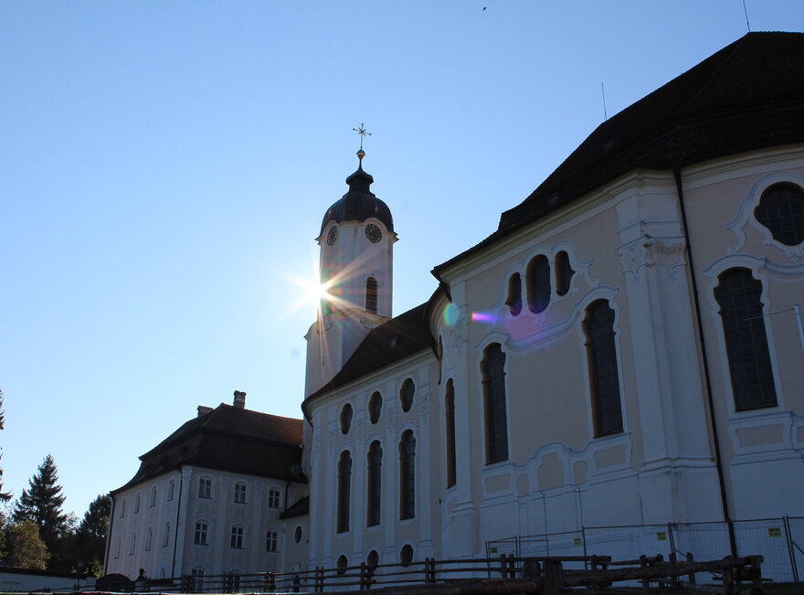  Das Bild zeigt die große weiße Wieskirche, mit einem hohen Kirchturm und einem Kreuz oben drauf. Die Sonne scheint hell auf die Kirche, sodass eine schöne und sommerliche Atmosphäre entsteht.