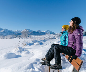 Das Bild zeigt zwei Frauen, die auf der Lehne einer tief verschneiten Holzbank sitzen. Sie tragen Winterkleidung, Handschuhe und Mützen, haben die Augen geschlossen und genießen die Sonnenstrahlen in ihrem Gesicht. Im Hintergrund sieht man die schneebedeckte Umgebung und die verschneiten Berge. Der Himmel ist blau. Die Sonne scheint.  