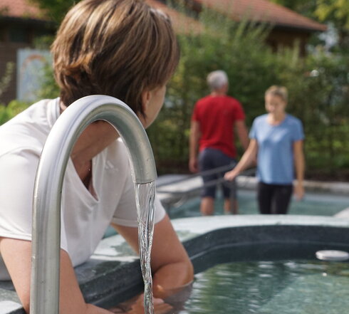 Das Bild zeigt im Vordergrund eine Frau, die ihre unbekleideten Unterarme in ein Armbecken taucht. Aus einem Wasserhahn davor läuft Wasser in das Armbecken. Im Hintergrund sieht man einen Mann und eine Frau in einem Wassertretbecken stehen. Die Wasserbecken sind mit Büschen und Bäumen im Hintergrund eingerahmt. Die Sonne scheint. 