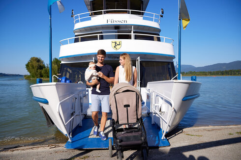 Das Bild fängt eine friedvolle Szene auf einem Schiff ein. Ein Mann und eine Frau stehen an Deck des Schiffs, das an einer Pier festgemacht ist und scheinen, als möchten sie von Borg gehen. Der Mann hält ein Baby in seinen Armen und die Frau steht neben ihm, einen Kinderwagen schiebend. Das Schiff ist weiß und blau und es weht eine Deutschland-Flagge im Wind, die am Schiff befestigt ist. Die Pier erstreckt sich ins türkisblaue Wasser des Forggensees. Im Hintergrund sind Bäume und Berge zu sehen. 