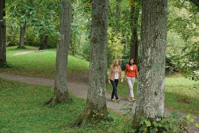 Das Bild zeigt zwei Frauen, die auf einen Feldweg laufen. Ringsum wachsen hohe Bäume. Der Boden ist mit Gras bewachsen. Die Sonne scheint. 