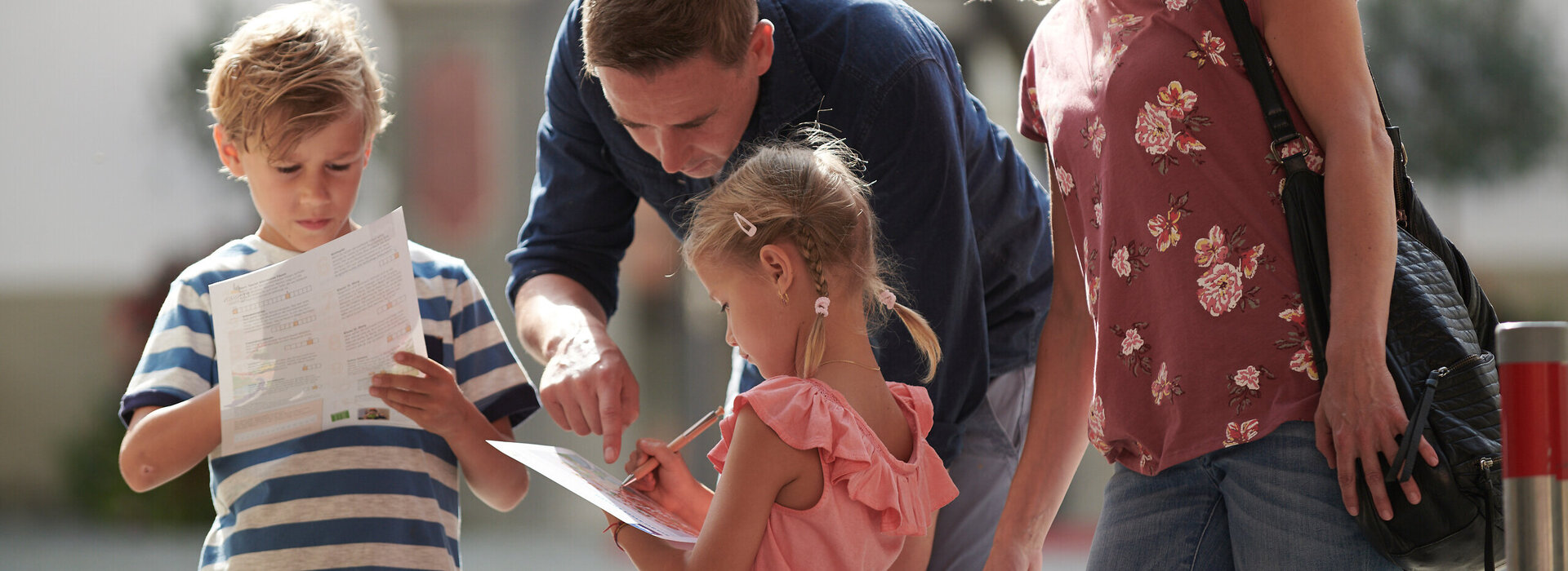 Das Bild zeigt eine vierköpfige Familie, bestehend aus einem Mann, einer Frau und zwei Kindern, die auf einer Straße stehen. Sie schauen auf ein Stadträtzel, welches Sie von der Tourist Information erhalten habe..