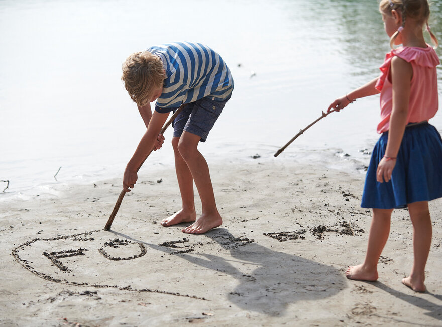 Das Bild zeigt eine ruhige Strandszene mit zwei jungen Kindern, die sich im Vordergrund befinden. Das Kind auf der linken Seite, das ein gestreiftes Hemd mit blauen und weißen Streifen und blaue Shorts trägt, beugt sich über die Erde, um einen Stock aufzuheben. Das Kind auf der rechten Seite, das ein rosa Shirt und einen blauen Rock trägt, hält einen Stock und weist damit auf das Wasser. Der sandige Strand ist mit kleinen Steinen und Kieseln gesprenkelt, was der Szene Textur verleiht. Das ruhige Wasser im Hintergrund vervollständigt die idyllische Kulisse.