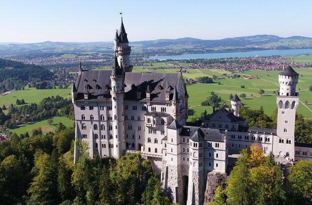 Das Bild zeigt das weiße Schloss Neuschwanstein, dass auf einer bewaldeten Anhöhe vor einer weiten Ebene liegt. Auf der Ebene sind Seen, die Gemeinde Schwangau und die Stadt Füssen zu sehen, die umgeben von Wäldern und grünen Wiesen sind. Das Schloss Neuschwanstein steht im Fokus des Fotos. Es besteht aus mehreren unterschiedlichen Gebäudeteilen, wie Türmen, Erkern und einem Torbau, die miteinander verbunden sind. Das Hauptgebäude des Schlosses ist 5-stöckig. Es hat ein graues Dach. Auf dem Hauptgebäude stehen eine überlebensgroße Statue sowie eine Löwenfigur. Der Himmel ist blau. Die Sonne scheint.
