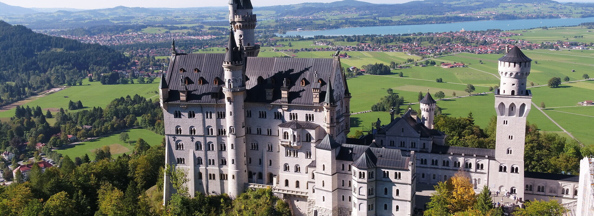 Das Bild zeigt das weiße Schloss Neuschwanstein, dass auf einer bewaldeten Anhöhe vor einer weiten Ebene liegt. Auf der Ebene sind Seen, die Gemeinde Schwangau und die Stadt Füssen zu sehen, die umgeben von Wäldern und grünen Wiesen sind. Das Schloss Neuschwanstein steht im Fokus des Fotos. Es besteht aus mehreren unterschiedlichen Gebäudeteilen, wie Türmen, Erkern und einem Torbau, die miteinander verbunden sind. Das Hauptgebäude des Schlosses ist 5-stöckig. Es hat ein graues Dach. Auf dem Hauptgebäude stehen eine überlebensgroße Statue sowie eine Löwenfigur. Der Himmel ist blau. Die Sonne scheint.