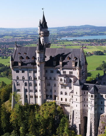 Das Bild zeigt das weiße Schloss Neuschwanstein, dass auf einer bewaldeten Anhöhe vor einer weiten Ebene liegt. Auf der Ebene sind Seen, die Gemeinde Schwangau und die Stadt Füssen zu sehen, die umgeben von Wäldern und grünen Wiesen sind. Das Schloss Neuschwanstein steht im Fokus des Fotos. Es besteht aus mehreren unterschiedlichen Gebäudeteilen, wie Türmen, Erkern und einem Torbau, die miteinander verbunden sind. Das Hauptgebäude des Schlosses ist 5-stöckig. Es hat ein graues Dach. Auf dem Hauptgebäude stehen eine überlebensgroße Statue sowie eine Löwenfigur. Der Himmel ist blau. Die Sonne scheint.