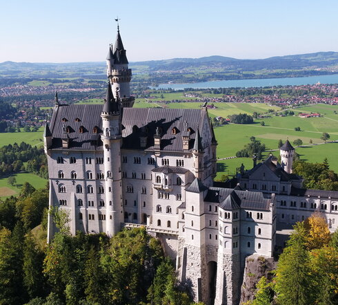 Das Bild zeigt das weiße Schloss Neuschwanstein, dass auf einer bewaldeten Anhöhe vor einer weiten Ebene liegt. Auf der Ebene sind Seen, die Gemeinde Schwangau und die Stadt Füssen zu sehen, die umgeben von Wäldern und grünen Wiesen sind. Das Schloss Neuschwanstein steht im Fokus des Fotos. Es besteht aus mehreren unterschiedlichen Gebäudeteilen, wie Türmen, Erkern und einem Torbau, die miteinander verbunden sind. Das Hauptgebäude des Schlosses ist 5-stöckig. Es hat ein graues Dach. Auf dem Hauptgebäude stehen eine überlebensgroße Statue sowie eine Löwenfigur. Der Himmel ist blau. Die Sonne scheint.
