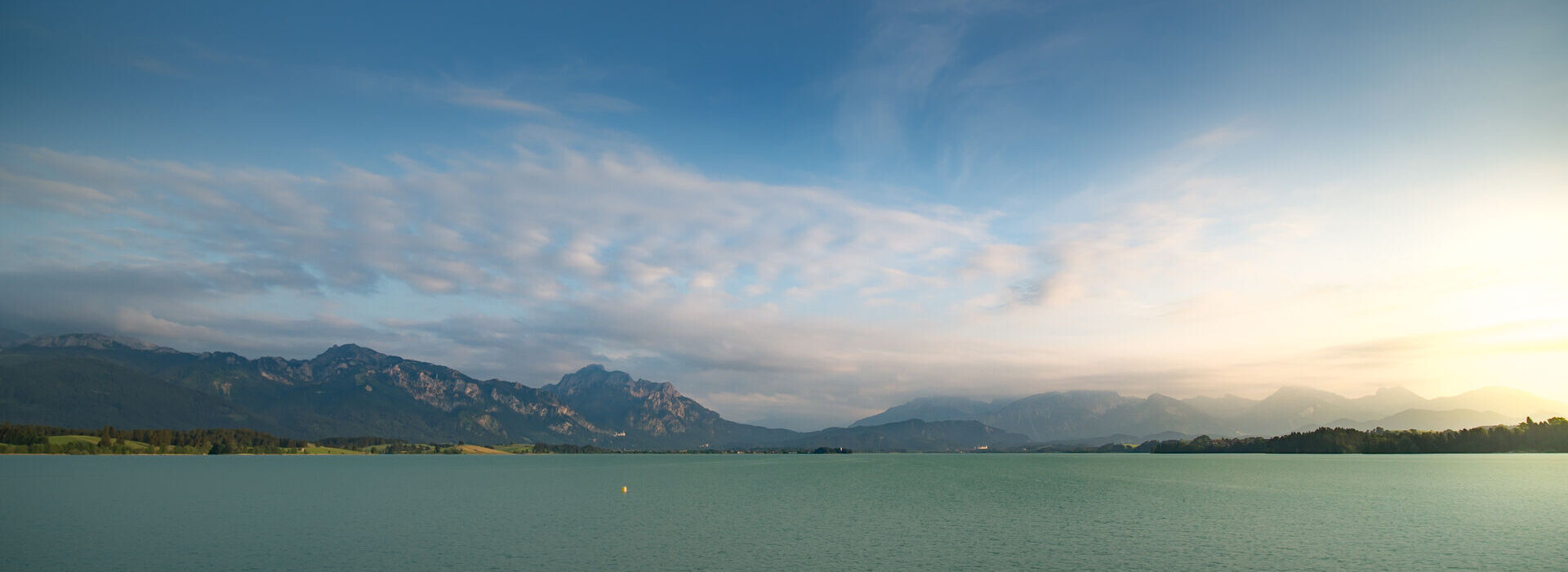 Das Bild zeigt eine idyllische Landschaft, die von einem weiten Gewässer dominiert wird, das sich zum Horizont hin erstreckt – dem Forggensee. Der türkise See ist ruhig und ungestört und spiegelt den klaren Himmel darüber wider. Der Himmel ist mit einigen verstreuten Wolken durchsetzt, die der Szene Tiefe und Dimension verleihen. In der Ferne erheben sich majestätische Berge, deren Gipfel den Himmel berühren. Die Berge, mit ihrem rauen Gelände, bieten einen scharfen Kontrast zum ruhigen Wasser. 