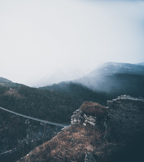 Das Bild zeigt eine Hängebrücke, die zwei Bergeplateaus miteinander verbindet. Auf der einen Anhöhe ist eine Burgruine zu sehen. Die Berge sind mit Wald bewachsen. Der Himmel ist rau. Nebel zieht auf. 