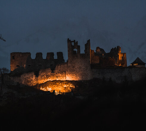 Das Bild zeigt eine beleuchtete Burgruine in der Abenddämmerung. Im Hintergrund erheben sich schneebedeckte Berge.  